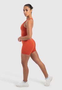 Orange träningsbodysuit med en figurnära design, som har tunna axelband och en sömlös, elastisk textur. Kombinerad med vita träningsskor.