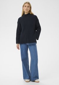 Femme se tenant debout, portant un pull à col roulé noir en maille côtelée, un jean bleu à jambes larges et des chaussures de couleur claire sur un fond blanc.