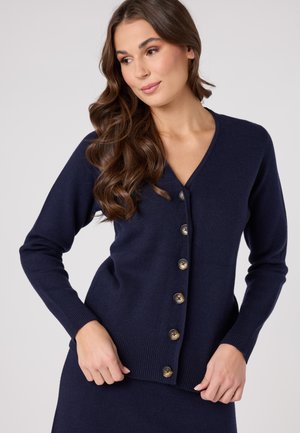 Cardigan blu navy con scollo a V, polsini a costine e sei grandi bottoni dorati sulla parte anteriore. Texture morbida, design aderente.