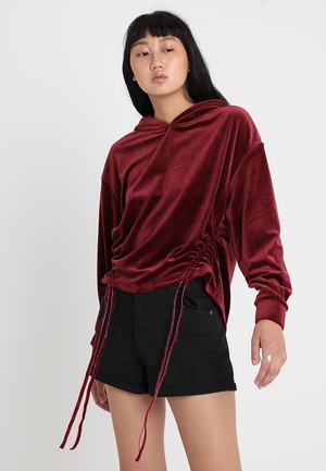 Hanorac din catifea burgundă cu tiv cu șnur și croiala supradimensionată. Asortat cu pantaloni scurți negri, având manșete răsucite și buzunare.