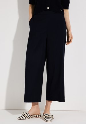 Frau trägt schwarze, weit geschnittene, verkürzte Hosen und schwarz-weiß gestreifte Slip-On-Schuhe mit Schleifendetail, steht vor weißem Hintergrund.