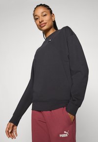 Mujer con cabello trenzado que lleva un sudadera negra de Puma y pantalones de chándal rojos de Puma, sonriendo con una mano en el bolsillo frente a un fondo liso.