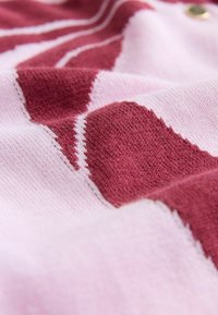 Pruhovaná pletenina v ružovej a burgundskej farbe s mäkkou textúrou a diagonálnym vzorom, doplnená kontrastným bielym prešívaním.