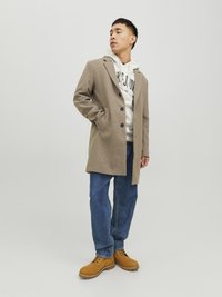 Beige Wollmischungsjacke mit Reverskragen, Frontknöpfen und Seitentaschen, über einem grauen Hoodie getragen, kombiniert mit blauen Jeans und braunen Stiefeln.