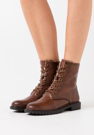 Botas de tornozelo em couro castanho com um acabamento suave, bico arredondado, design com atacadores e sola de borracha texturizada. Os destaques incluem ilhoses de metal e costura.