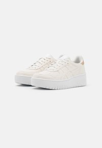 Un paio di sneaker basse color avorio con suole bianche spesse, design con lacci e sottili strisce del marchio sui lati, su uno sfondo neutro.