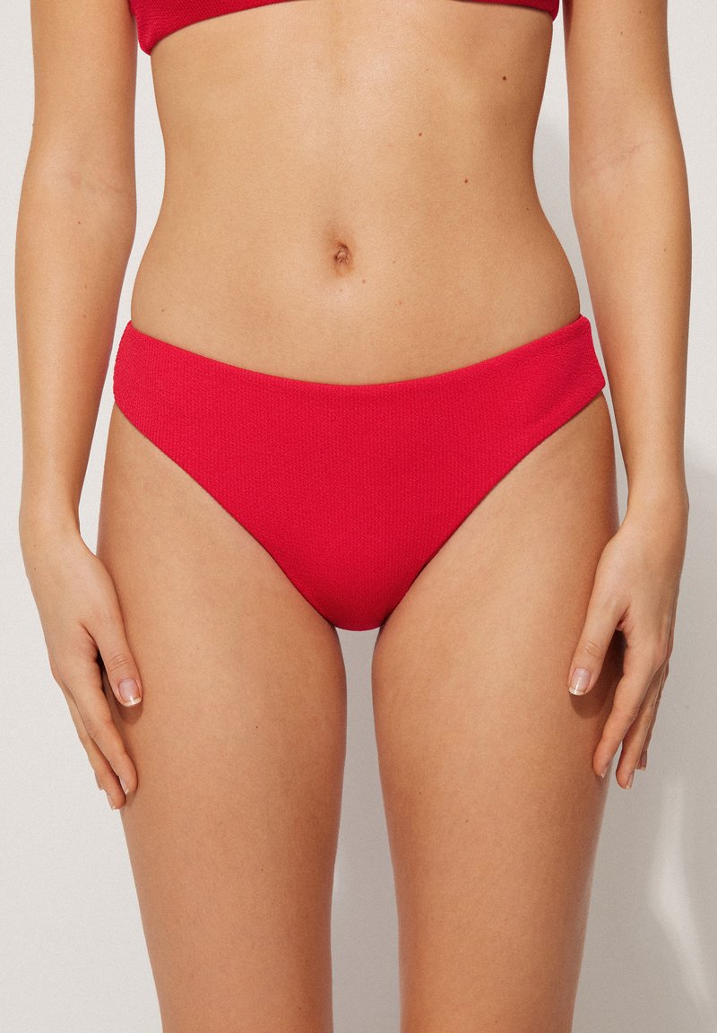 Bas de bikini rouge en tissu texturé avec une taille mi-haute. Offre une couverture minimale et une finition lisse. Pas de matériel visible.