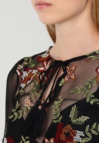 Top nero trasparente con intricati ricami floreali in rosso, verde e bianco. Presenta un collo con lacci e frange.