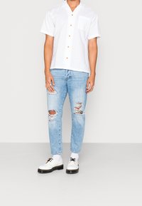 Camicia bianca a maniche corte con bottoni e taschino, abbinata a jeans chiari strappati e scarpe bianche con suola nera.