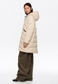 Abrigo beige acolchado con capucha y bolsillo lateral con cremallera, con un ajuste ligeramente oversize y una textura suave. Combinado con pantalones sueltos de color verde oliva.