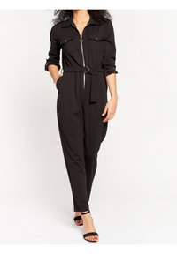 Black jumpsuit with a zip front, long sleeves, two chest pockets, a belted waist, and tapered legs. Satin-like fabric with a smooth texture.