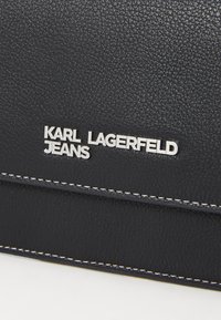 Juodas tekstūruotas oda su baltais siūlais; priekyje yra sidabrinis logotipas su užrašu "KARL LAGERFELD JEANS" įspaustas.