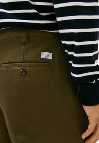 Olijfgroen broek met een achterzak voorzien van een knoop en een label. Gestreepte marineblauwe en witte trui gedeeltelijk zichtbaar aan de rechterkant.