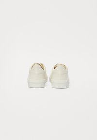 Baskets de couleur crème avec une finition en cuir lisse, un talon rembourré et une semelle en caoutchouc texturée, présentant un dos arrondi et un design minimal.