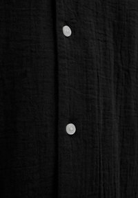 Camisa negra de botones con un tejido texturizado, botones blancos y un diseño sencillo con un acabado arrugado sutil.