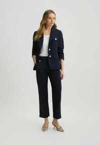 Navy blazer with silver buttons and side pockets, paired with navy trousers and patterned heels. The blazer has a tailored fit and rolled sleeves.