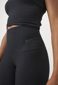 Melni riboti legingi ar augstu jostasdaļu un nelielu melnu Nike logotipu sānos. Audumam ir gluda tekstūra.