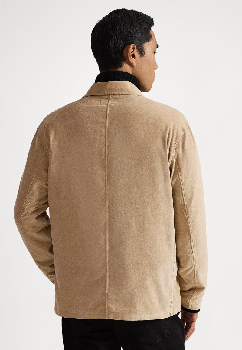 Mann trägt eine beige Cordjacke über einem schwarzen Rollkragenpullover, von hinten vor einem schlichten weißen Hintergrund gezeigt.