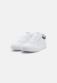 Bílé tenisky s hladkým koženým svrškem, šněrovacím designem, polstrovaným límcem a texturovanou gumovou podrážkou. Obsahují nenápadné logo na jazyku.