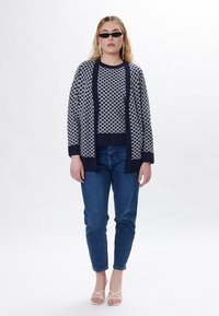 Cardigan lavorato a maglia a righe blu e bianche sopra una maglietta bianca, abbinato a jeans blu e sandali bianchi con cinturini. Tessuto morbido, vestibilità rilassata.