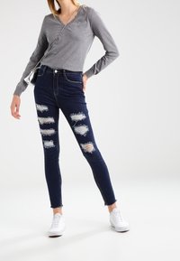 Femme portant un pull gris à col en V, un jean skinny déchiré bleu foncé, des baskets blanches et un petit sac en bandoulière argenté sur fond blanc.