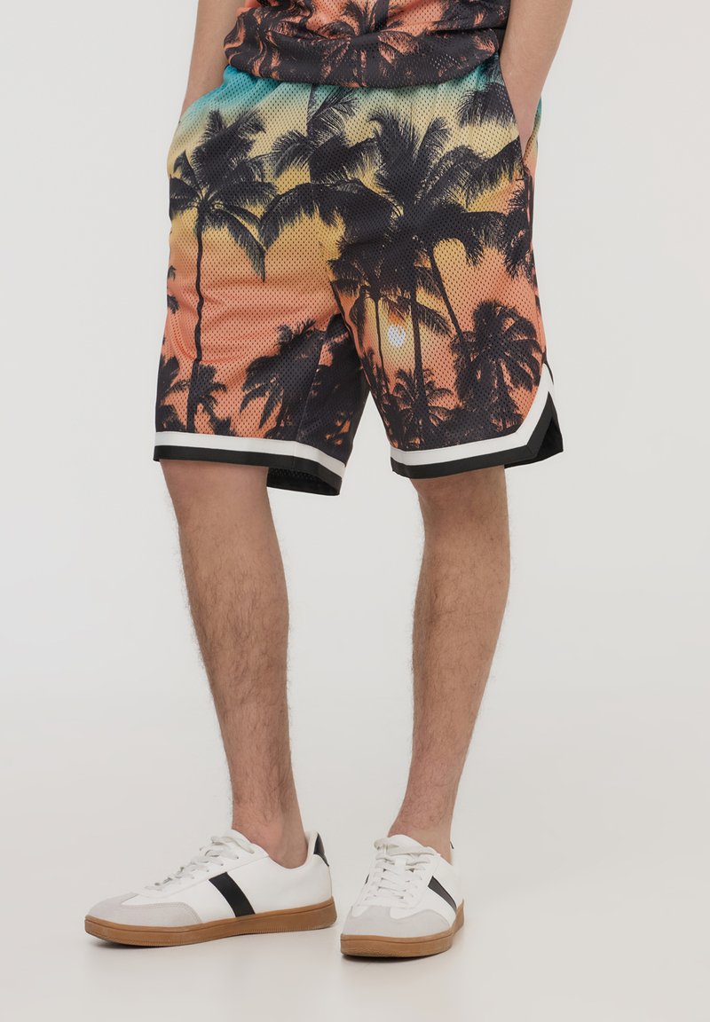 Pantaloni corti a rete multicolori con design a tramonto con palme, bordo nero e tasche laterali. Abbinati a sneakers bianche e dettagli neri.