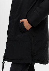 Schwarze Pufferjacke mit Reißverschluss, strukturiertem Material, seitlichen Taschen und verstellbarem Saum. Hand in die Tasche gelegt.