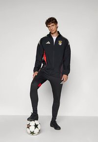 Black Adidas tracksuit with red accents, featuring a zip-up jacket and fitted pants. The model stands on a soccer ball.