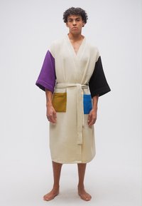 Νεαρός άνδρας στέκεται ξυπόλητος, φορώντας ένα ρόμπα από βαμβακερό ύφασμα βάφλ σε απαλό κρεμ χρώμα, με μανίκια σε μωβ και μαύρο και μπροστινές τσέπες σε μουσταρδί και μπλε.