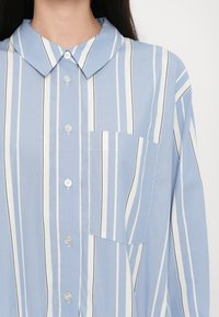 Chemise bleu clair boutonnée avec des rayures verticales blanches et noires, poche sur la poitrine gauche et col arrondi.