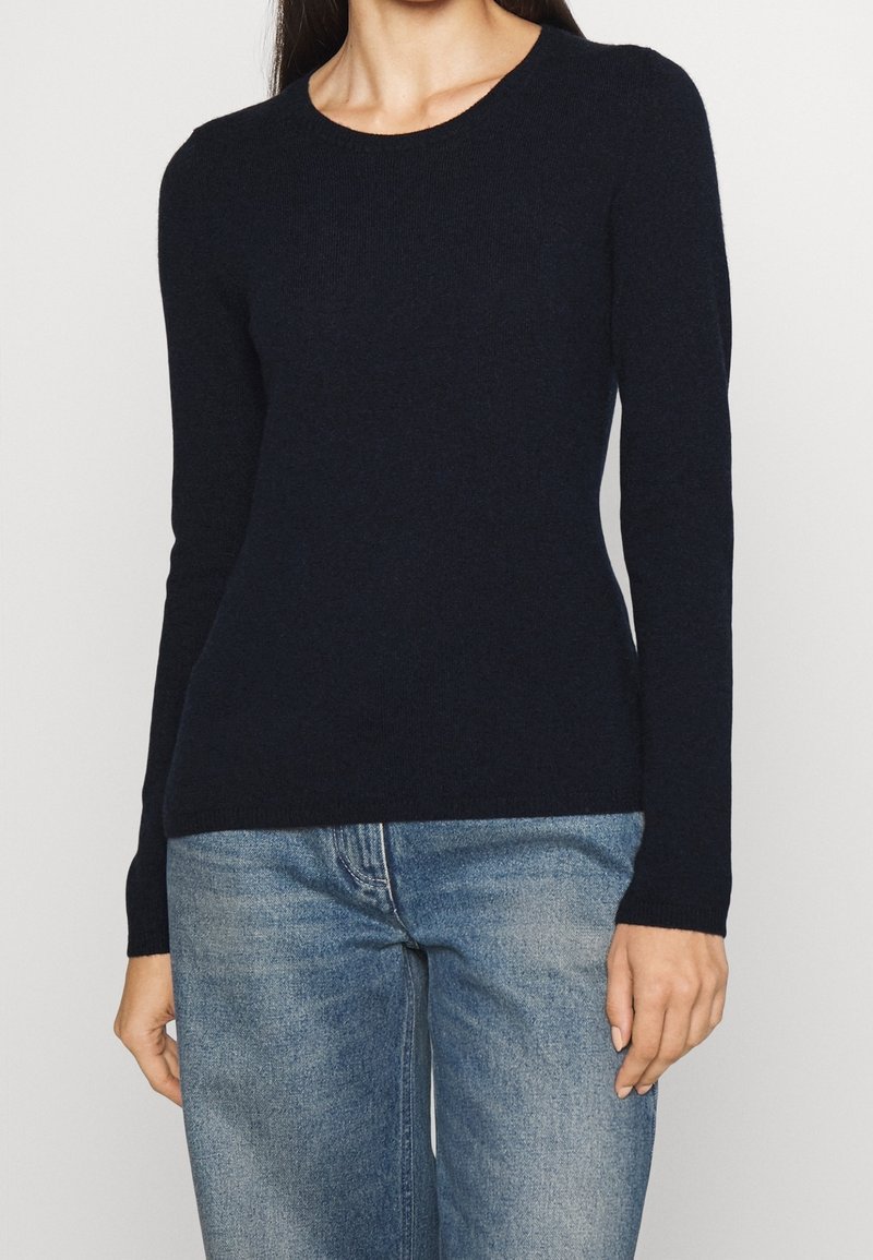 Femme portant un pull noir ajusté à manches longues et un jean denim bleu clair sur fond uni.