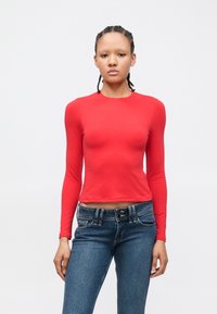 Ung person med flettet hår iført en tettsittende rød genser med lange ermer og blå jeans, stående foran en enkel lys bakgrunn.