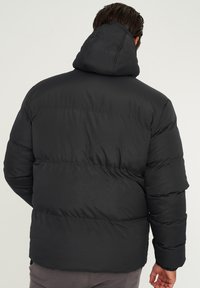Schwarze Steppjacke mit Kapuze, ausgestattet mit gesteppten Abschnitten und elastischen Bündchen. Glatte, matte Oberfläche mit enger Passform.