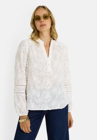 Blusa blanca bordada con cuello mandarín, tapeta de botones, patrones florales y mangas largas con detalles de encaje. Combinada con pantalones vaqueros oscuros.