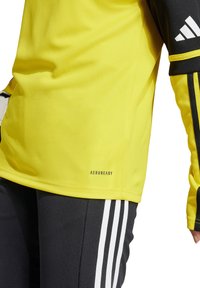 Gelbes Sportoberteil mit feuchtigkeitsregulierender AEROREADY-Technologie, schwarzen Akzenten und kontrastierenden schwarzen Streifen an den Ärmeln und Hosen.