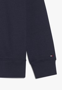 Ciemnogranatowy rękaw bluzy z prążkowanym teksturą i subtelnym detalem logo z czerwonymi, białymi i niebieskimi akcentami. Miękki materiał, klasyczny design.