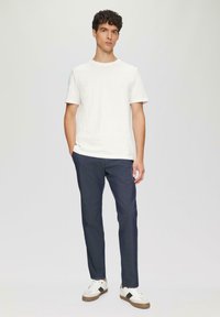 Weißes T-Shirt mit Rundhalsausschnitt, dunkelblaue Hose mit dezentem Muster und sportliche weiße Schuhe mit braunen Akzenten, stehen vor einem neutralen Hintergrund.