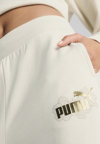 Kremowe dresy z żebrowanym pasem w talii mają złote i czarne logo "PUMA" oraz kwiatowy wzór po lewej stronie kieszeni.