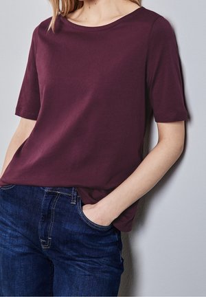 Frau trägt ein schlichtes weinrotes Kurzarmshirt und dunkelblaue Jeans, mit einer Hand in der Tasche, vor einer hellgrauen Wand.