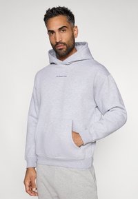 Ανοιχτό γκρι φούτερ από μαλακό ύφασμα, με καγκουρό τσέπη, κουκούλα με κορδόνια και μικρό κεντητό λογότυπο στο στήθος.