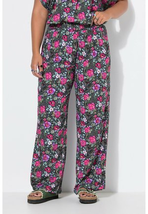 Person iført løse bukser med blomsterprint i pink, lilla og hvid, kombineret med grønne åbne sandaler og ringe på fingrene.