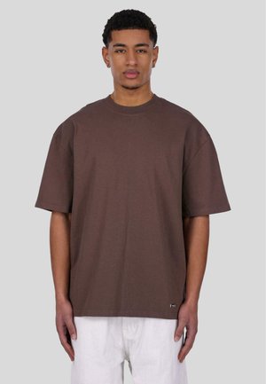 Brown cotton T-shirt with short sleeves, a crew neck, and a relaxed fit, paired with white pants, featuring a small logo at the hem.