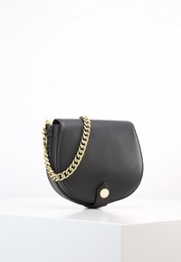 Borsa a tracolla in pelle nera con forma curvata, tracolla in catena dorata e chiusura rotonda. Texture liscia con elementi minimi.