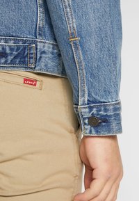 Chaqueta de mezclilla azul con puños abotonados; pantalones color beige con una etiqueta de Levi's. Presenta costuras naranjas y un acabado desgastado en la chaqueta.