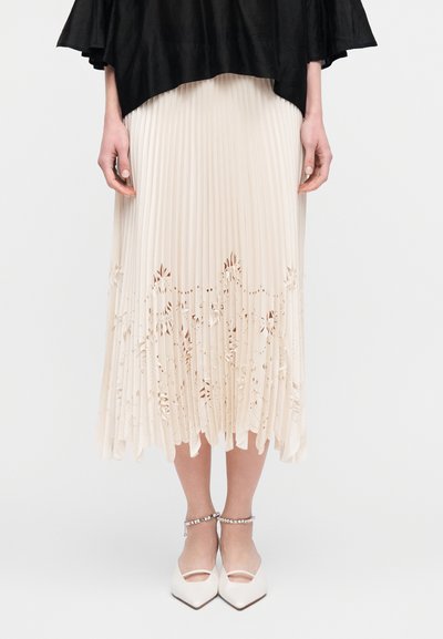 Φούστα από ελεφαντόδοντο με πιέτες και λεπτομέρειες με λουλουδάτα κοψίματα στο τελείωμα, συνδυασμένη με λευκές μυτερές γόβες με λουράκι στον αστράγαλο. Ορατό είναι επίσης το μαύρο τοπ.