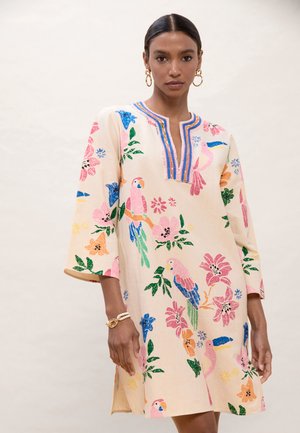 Mujer con un vestido beige hasta la rodilla con coloridos bordados de aves tropicales y flores, mangas tres cuartos y escote con muesca.