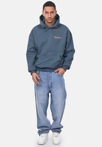 Sweat-shirt à capuche gris foncé avec poche avant et logo rose, associé à un jean baggy bleu clair et des baskets blanches.