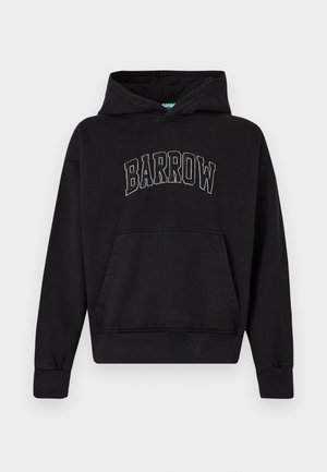 Mężczyzna ubrany w czarną bluzę z kapturem z napisem "BARROW", jeansowe szorty z łańcuszkiem, białe skarpetki, czarne trampki i okulary przeciwsłoneczne, stoi ze rękami w kieszeniach.