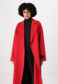 Roter Wollmantel mit doppelter Knopfleiste, Reverskragen und seitlichen Taschen, kombiniert mit einem schwarzen Rollkragenpullover und einer Hose.