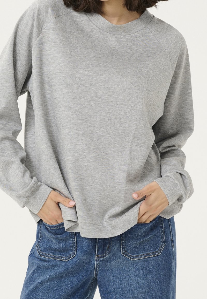 Personne portant une chemise à manches longues gris clair et un jean bleu en denim, les mains dans les poches avant.